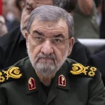 Older man in a dark green military uniform with gold epaulets and red collar insignia, looking at the camera, wearing glasses.